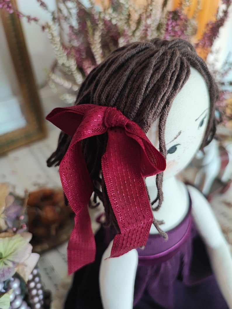 May include: A handmade doll with brown yarn hair and a burgundy ribbon tied in a bow. The doll is wearing a purple dress. The doll's face is painted with delicate features, including small eyes and freckles.