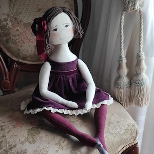 May include: A handmade cloth doll with brown yarn hair and a burgundy ribbon. The doll wears a plum-colored dress with a white lace trim and matching tights. It is seated on a vintage chair with floral upholstery.