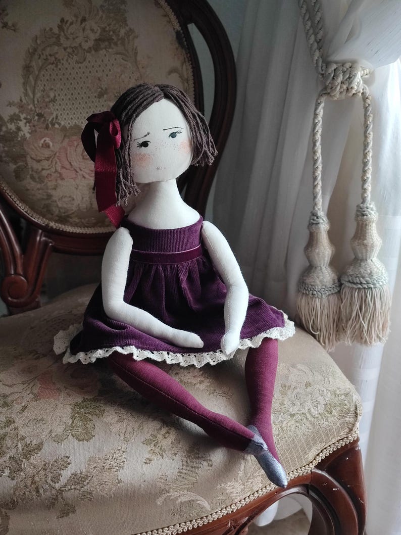 May include: A handmade cloth doll with brown yarn hair and a burgundy ribbon. The doll wears a plum-colored dress with a white lace trim and matching tights. It is seated on a vintage chair with floral upholstery.