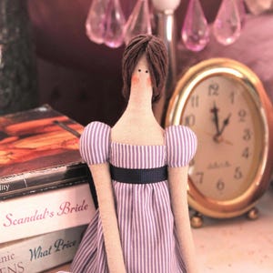 May include: A handmade doll with brown hair, wearing a purple and white striped dress with a black sash. The doll is sitting on a table with a book and a clock in the background.