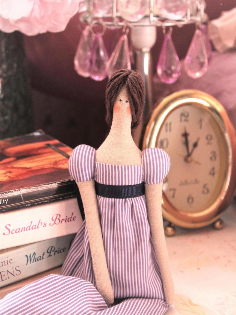 May include: A handmade doll with brown hair, wearing a purple and white striped dress with a black sash. The doll is sitting on a table with a book and a clock in the background.
