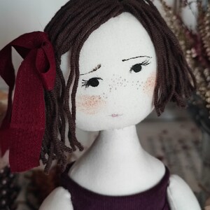 May include: Handmade cloth doll with brown yarn hair and a burgundy ribbon. The doll has a painted face with freckles, rosy cheeks, and a purple dress. A unique, handcrafted item.