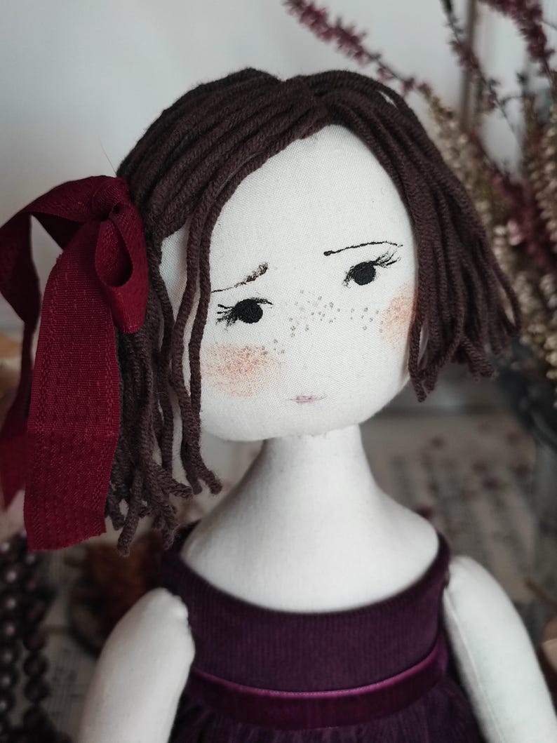 May include: Handmade cloth doll with brown yarn hair and a burgundy ribbon. The doll has a painted face with freckles, rosy cheeks, and a purple dress. A unique, handcrafted item.