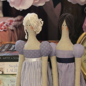 May include: Two handmade fabric dolls with long limbs and striped dresses. The doll on the left has blonde hair and a purple dress with white polka dots. The doll on the right has dark brown hair and a purple and white striped dress.