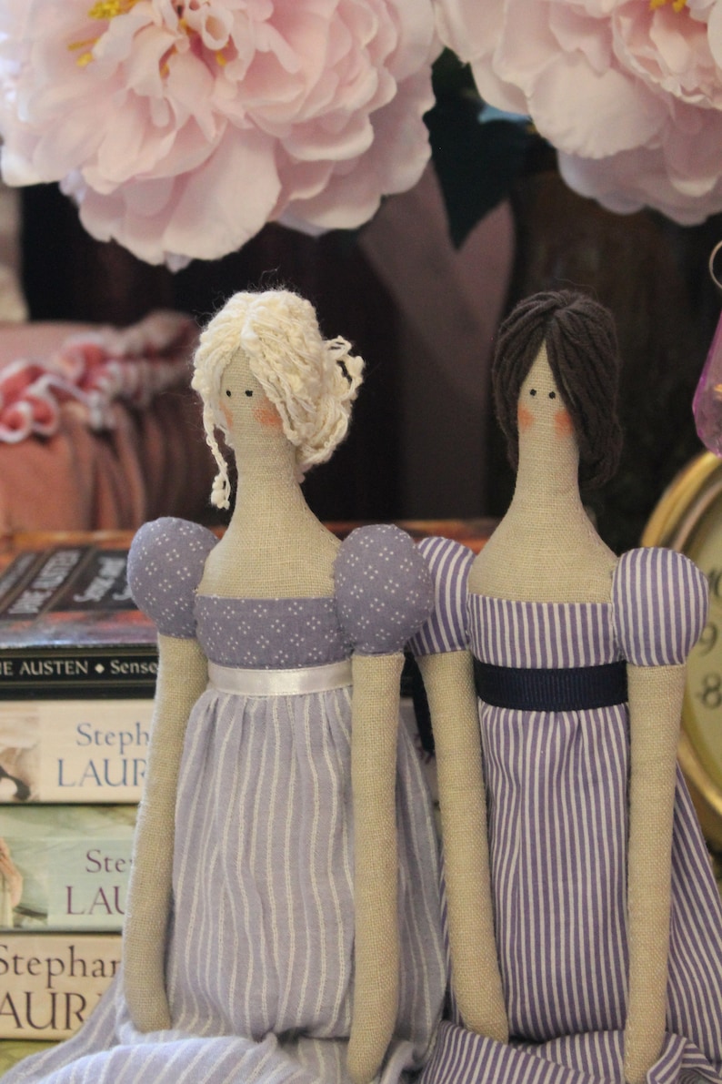 May include: Two handmade fabric dolls with long limbs and striped dresses. The doll on the left has blonde hair and a purple dress with white polka dots. The doll on the right has dark brown hair and a purple and white striped dress.