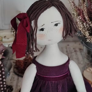 May include: A handmade cloth doll with brown yarn hair and a burgundy ribbon. The doll has a white face with freckles and rosy cheeks, wearing a dark purple corduroy dress with a velvet ribbon.