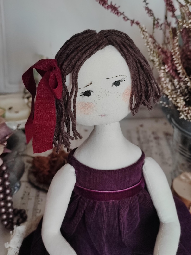 May include: A handmade cloth doll with brown yarn hair and a burgundy ribbon. The doll has a white face with freckles and rosy cheeks, wearing a dark purple corduroy dress with a velvet ribbon.