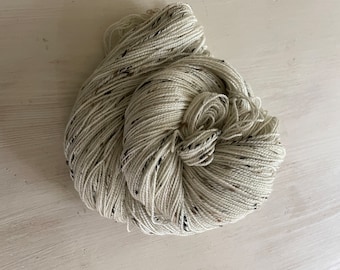 May include: A skein of off-white yarn with flecks of black and brown. The yarn is tightly wound, with a portion of the skein unwound, revealing the texture of the fibers. Ideal for knitting, crocheting, or other fiber arts projects.