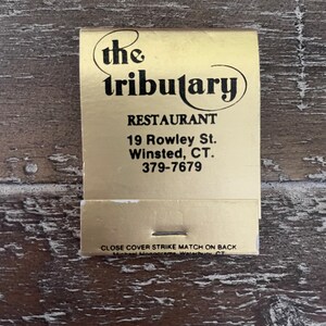 May include: A gold matchbook with black text that reads "the tributary RESTAURANT 19 Rowley St. Winsted, CT. 379-7679 CLOSE COVER STRIKE MATCH ON BACK Michael Monograms Waterbury CT"