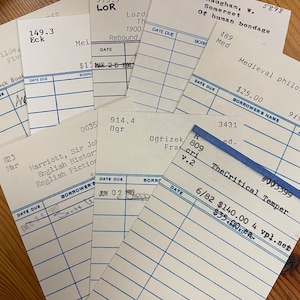 May include: Close-up of several library card pockets with blue lines. The cards are filled out with titles, authors, and due dates. Some of the cards have handwritten notes.