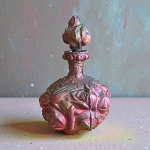 May include: A vintage-style perfume bottle with a rose motif. The bottle is a metallic pink and bronze color, with a bulbous body and a slender neck. The stopper is a detailed, ornate design. The bottle is likely made of metal or glass.