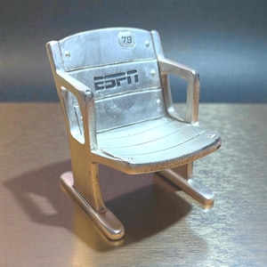 May include: A silver-colored miniature stadium seat with the ESPN logo and the number 79. The seat has armrests and a slatted seat design, resembling a vintage stadium chair. The chair is made of metal.