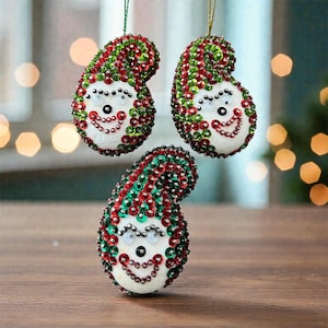 May include: Three Christmas ornaments shaped like smiling elves. Each ornament is white with red, green, and silver sequins. The ornaments have black eyes and a smiling mouth. They are hanging from green and gold strings.