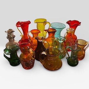 May include: A collection of various glass pitchers in vibrant colors including red, yellow, orange, green, and amber. Each pitcher features a unique crackle glass texture, a handle, and a ruffled or flared rim, showcasing diverse shapes and sizes.