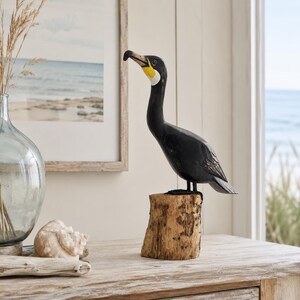 Puede incluir: Una figura de cormor&aacute;n tallada a mano en madera con cuerpo negro, una mancha blanca en la garganta y detalles amarillos en el pico. El ave est&aacute; posada sobre una base de tronco de madera natural y texturizada, mostrando vetas y detalles.
