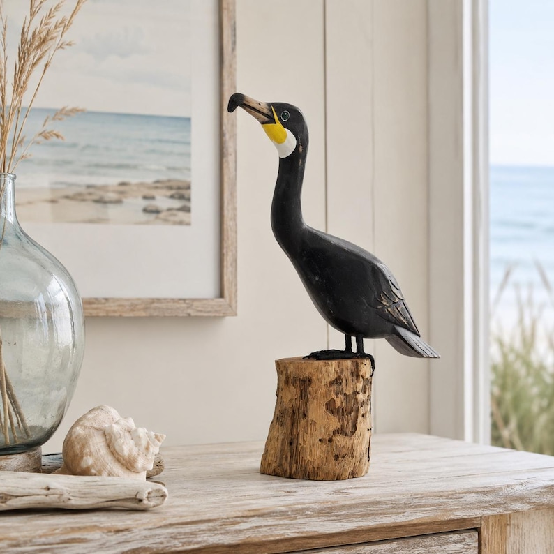 Puede incluir: Una figura de cormor&aacute;n tallada a mano en madera con cuerpo negro, una mancha blanca en la garganta y detalles amarillos en el pico. El ave est&aacute; posada sobre una base de tronco de madera natural y texturizada, mostrando vetas y detalles.