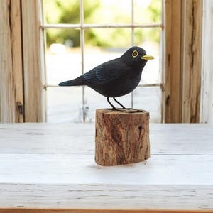 Könnte beinhalten: Eine schwarze Holzvogelfigur, die auf einem kleinen Holzstumpf sitzt. Der Vogel hat einen gelben Schnabel und schwarze Augen.