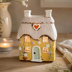 May include: A ceramic cottage-shaped tea light holder with a yellow facade, a white thatched roof, and a central heart cutout. The exterior features painted floral vines, four square windows, and a light blue arched door with a black knob.
