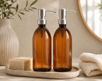 May include: Two amber glass soap dispensers with silver pump tops. The dispensers sit on a light-coloured tray with a bar of soap. A white towel and a decorative vase are also in the background. The dispensers are ideal for a bathroom or kitchen.