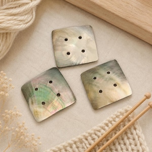May include: Three square buttons made of iridescent mother-of-pearl shell. Each button features a smooth, polished surface with a natural pearlescent sheen displaying subtle green, pink, and grey tones. Every button has four holes.