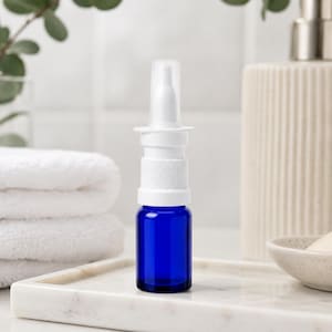May include: A cobalt blue glass nasal spray bottle with a white applicator. The bottle is on a marble tray, with a stack of white towels and a decorative dispenser in the background. The bottle is empty.