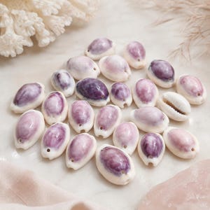 May include: A collection of small, oval-shaped cowrie shells, each featuring a smooth, white exterior with a prominent, deep purple to light lavender centre. Each shell has a small, circular hole drilled at one end, indicating their use as beads.