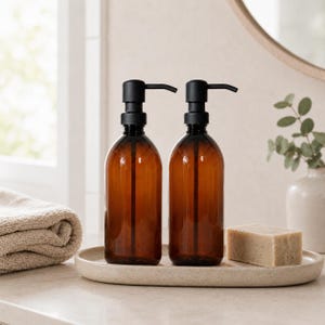 Puede incluir: Dos botellas dispensadoras de jabón recargables de color ámbar con tapas de bomba negras. Las botellas se encuentran en una bandeja ovalada moteada con una pastilla de jabón. Una toalla enrollada y un jarrón con vegetación están en el fondo.