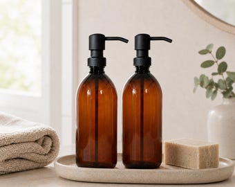 May include: Two amber-coloured, refillable soap dispenser bottles with black pump tops. The bottles sit on a speckled, oval tray with a bar of soap. A rolled towel and a vase with greenery are in the background.