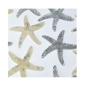Könnte beinhalten: Eine Sammlung von schillernden Seesternen in Silber und Weiß. Die Seesterne sind aus Harz und haben eine glitzernde Oberfläche.