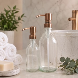 Op de afbeelding: Twee doorzichtige glazen zeepdispensers met koperkleurige pompen. De ene dispenser is groter dan de andere. De dispensers staan op een marmeren oppervlak, met witte handdoeken en zeep op de achtergrond. De dispensers zijn leeg.