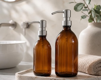 May include: Two amber glass soap dispensers with brushed metal pump heads. The bottles feature a vertical ribbed texture and are placed side by side. The dispensers have a cylindrical shape with a rounded base and a long, slender neck leading to the pump.