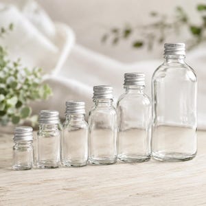 May include: A set of clear glass bottles with silver metal lids, displayed in ascending order of size. The smallest bottle is on the left, the largest on the right. The background is a soft, blurred white and green, with a wooden surface.