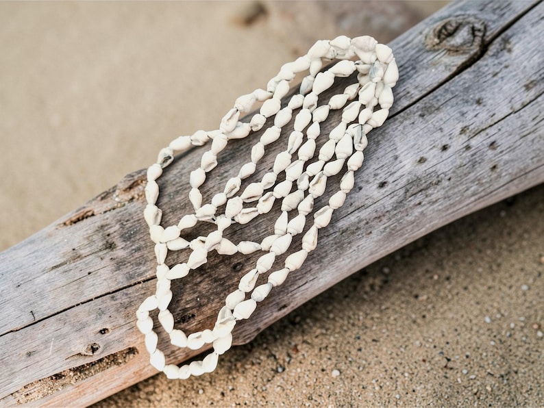 Puede incluir: Un collar de conchas blancas con m&uacute;ltiples hilos, apoyado sobre un trozo de madera flotante desgastada. El collar est&aacute; compuesto por peque&ntilde;as conchas de forma ovalada, creando una est&eacute;tica texturizada y natural. El fondo es una playa arenosa.