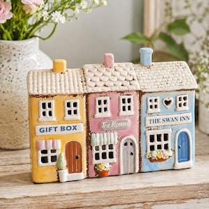 May include: A ceramic decorative building figurine featuring three connected storefronts: a yellow Gift Box, a pink Tea Rooms, and a blue The Swan Inn. Each section has a unique roof, small windows, and a door, with yellow, pink, and blue chimneys.