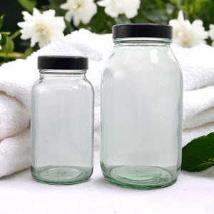 Puede incluir: Dos frascos de vidrio transparente con tapas negras. Los frascos son de diferentes tamaños, el más grande a la derecha. El fondo incluye toallas blancas, flores blancas y follaje verde. Los frascos están vacíos.