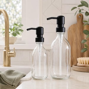 May include: Two clear glass soap dispensers with black pump tops. The dispensers are different sizes and sit on a white countertop. A gold tap and a green towel are in the background. The dispensers are for kitchen or bathroom use.