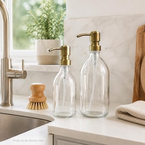 May include: Two clear glass soap dispensers with gold-coloured pumps. The dispensers are different sizes and sit on a white countertop. A wooden dish brush and a folded beige towel are also visible. The text "Perfect by the Kitchen Sink" is at the bottom.
