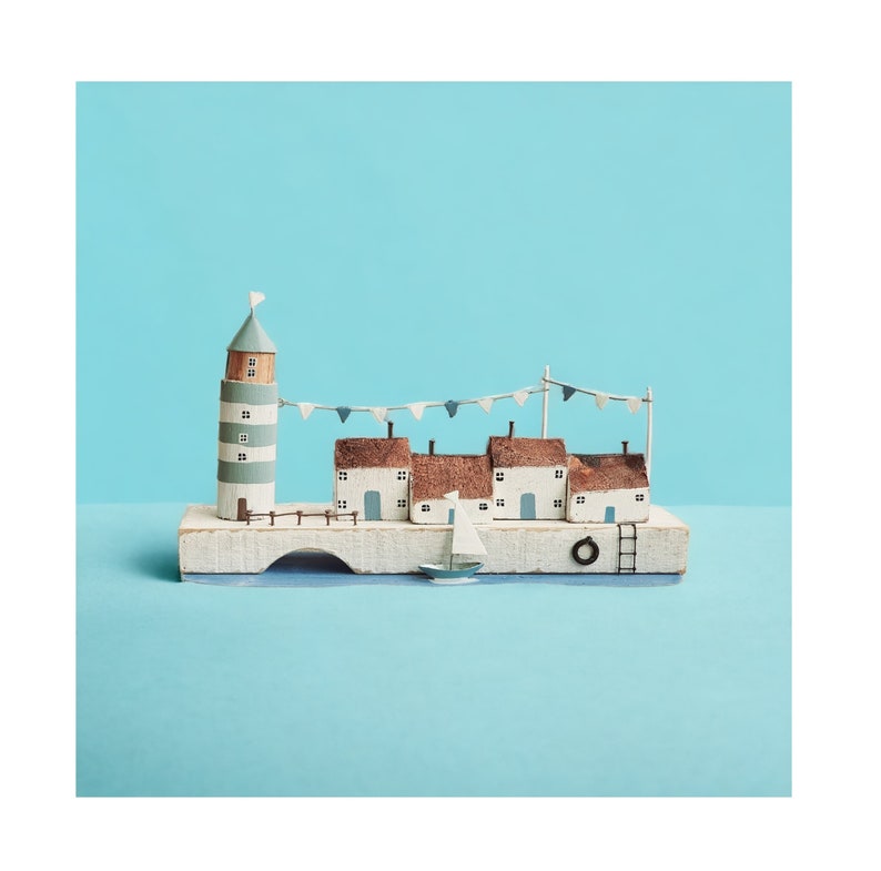 Pu&ograve; includere: Un villaggio costiero in miniatura di legno con un faro, case, un ponte e una barca a vela. Le case hanno pareti bianche, tetti marroni e dettagli blu. Il faro &egrave; a strisce blu e bianche. Il ponte &egrave; bianco con un arco blu. La barca a vela &egrave; bianca con una vela blu. Il villaggio &egrave; posto su una base di legno.