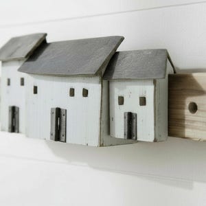 以下が含まれることがあります： グレーの屋根が付いた3つの小さな白い家が特徴の木製の壁の装飾。各家には暗い色のドアと正方形の窓があります。家は右側の木製のブロックに取り付けられています。