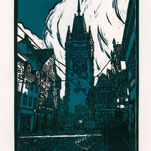 May include: A teal blue woodcut print of a street scene in Freiburg, Germany. The print features a tall clock tower with a pointed roof and a person walking in the foreground. The text "Freiburg, Germany" and "Miyo 2016" are visible in the lower right and left corners of the print.