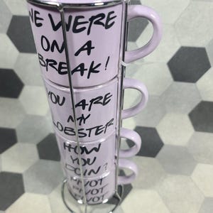 May include: A stack of four lavender mugs with black text, held in a silver metal rack. The mugs have phrases from a popular TV show, including "WE WERE ON A BREAK!" and "YOU ARE MY LOBSTER."