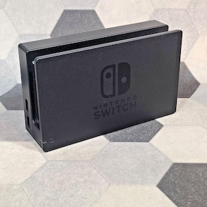 May include: A black Nintendo Switch dock, featuring the Nintendo Switch logo and text. The dock is rectangular with a slot for the console and ports on the side. The background is a hexagonal tile pattern.