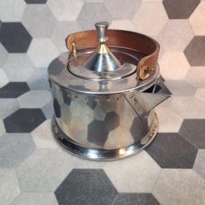 May include: A silver teapot with a hammered finish and a brown leather handle. The teapot features a conical lid with a silver and gold finial. The spout is triangular. The teapot sits on a round base.