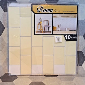 May include: A package of decorative wall tiles with a light yellow and white rectangular pattern. The package includes a label with the text "Room Decor" and "Decorative Wall Tiles", and the number "10 sheets".