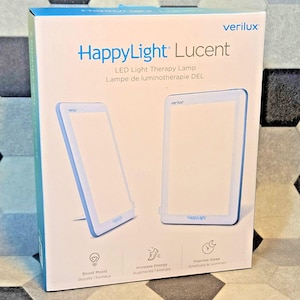 Könnte beinhalten: Eine weiße und blaue Box für eine HappyLight Lucent LED-Lichttherapielampe. Die Box zeigt zwei Bilder der Lampe mit Text, der darauf hinweist, dass sie die Stimmung aufhellen, die Energie steigern und den Schlaf verbessern kann. Die Marke "verilux" ist sichtbar.