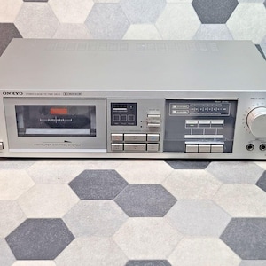 Puede incluir: Una pletina de casete estéreo Onkyo plateada con sistema de control por ordenador, compartimento central para casetes, múltiples botones de control, pantalla de nivel de pico, un gran mando de volumen y dos tomas de entrada de micrófono.