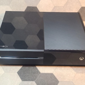 Könnte beinhalten: Eine schwarze Xbox One Konsole mit einem geometrischen Muster auf der linken Seite und einer strukturierten Oberfläche auf der rechten Seite. Das Xbox-Logo ist sichtbar.