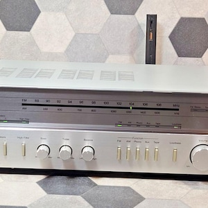 Könnte beinhalten: Silberner Vintage-Stereo-Receiver mit gebürsteter Metallfront. Er verfügt über ein Tuner-Zifferblatt, verschiedene Bedienknöpfe und einen Kopfhöreranschluss. Der obere Teil hat Lüftungsschlitze. Eine schwarze Fernbedienung steht hinter dem Receiver.