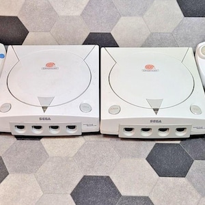 Może przedstawiać: Dwie białe konsole do gier Sega Dreamcast oraz cztery pasujące białe kontrolery przewodowe. Każdy kontroler posiada pad kierunkowy, cztery kolorowe przyciski akcji oraz gniazdo na kartę pamięci. Pojedyncza karta pamięci znajduje się w lewym dolnym rogu.