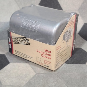 May include: A silver-colored Red Dot Code Keeper wet location cover in its original packaging. The box features red and black text, including the words Wet Location Cover, Code Keeper, and product features. The cover is made of cast aluminum.